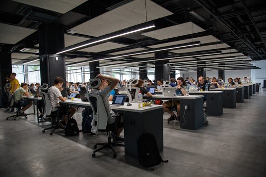 Office workers working on laptops inside an office room with many rows of desks.